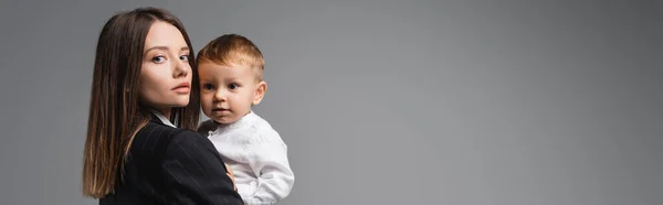 Молода бізнес-леді тримає малюка і дивиться на камеру ізольовано на сірому, банер — стокове фото