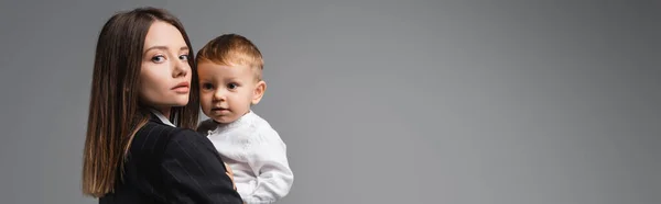 Young businesswoman holding toddler kid and looking at camera isolated on grey, banner — Stock Photo