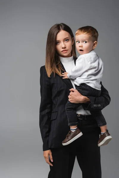 Mujer en traje negro sosteniendo hijo sorprendido aislado en gris - foto de stock