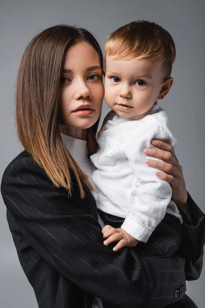 Mujer de negocios en chaqueta negra abrazando al niño y mirando a la cámara aislada en gris - foto de stock