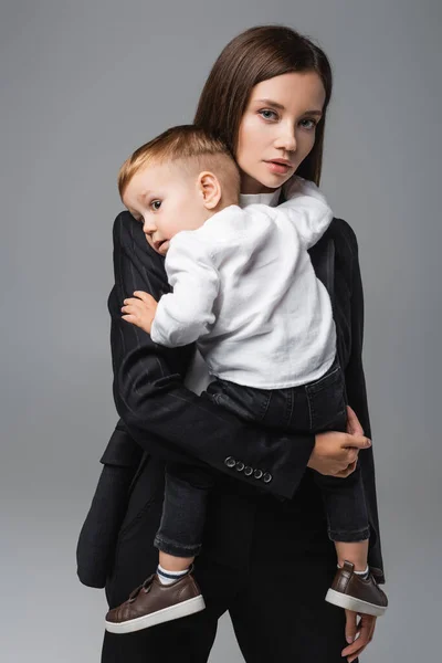 Young businesswoman looking at camera while holding son isolated on grey — стоковое фото