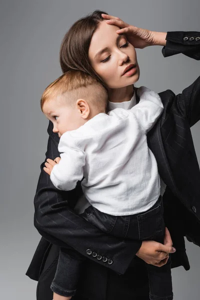 Mujer con los ojos cerrados tocando la cabeza mientras sostiene bebé niño aislado en gris - foto de stock