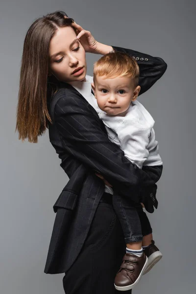 Jeune femme fixant les cheveux tout en tenant son fils grimaçant à la caméra isolée sur gris — Photo de stock