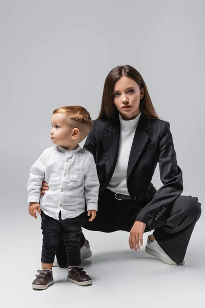 Young woman in black suit looking at camera while posing near toddler son on grey — Stock Photo