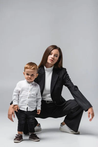 Молода жінка в чорному костюмі позує біля маленького хлопчика на сірому — стокове фото