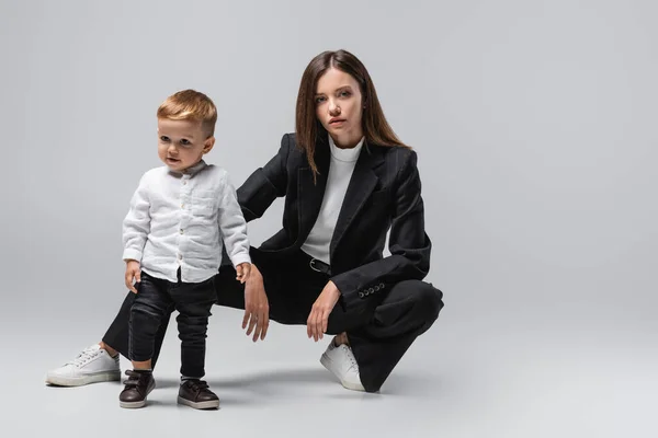 Mujer de negocios en traje negro posando cerca de pequeño hijo de pie sobre gris - foto de stock