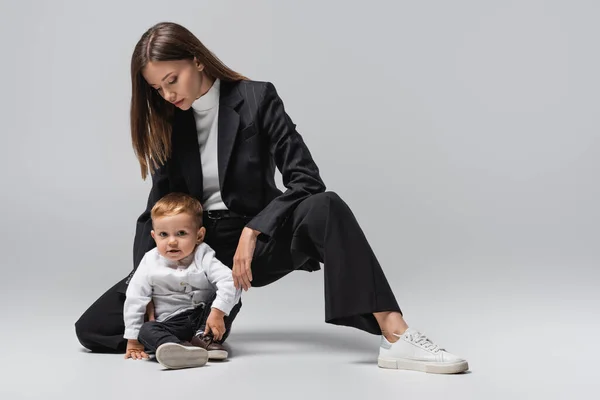 Niño mirando a la cámara mientras está sentado cerca de mamá en traje negro en gris - foto de stock