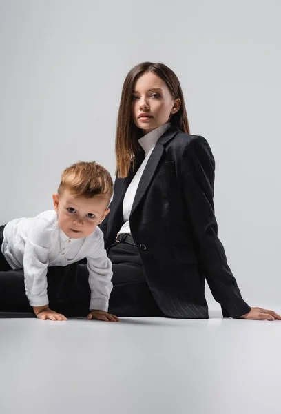 Woman in black suit looking at camera while sitting near crawling son on grey — Foto stock
