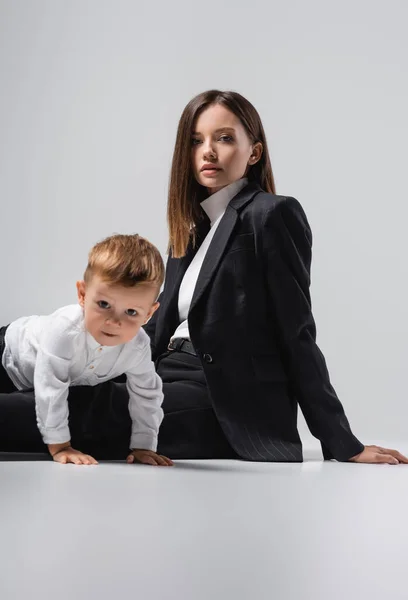 Mujer en traje negro mirando a la cámara mientras está sentado cerca de gatear hijo en gris - foto de stock