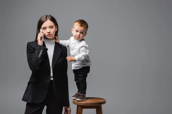 Woman in black suit talking on smartphone near little son standing on high stool isolated on grey — Foto stock