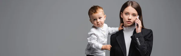 Mujer en blazer negro hablando en el teléfono móvil cerca de pequeño hijo aislado en gris, bandera - foto de stock