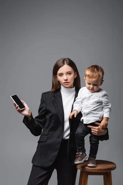 Жінка тримає смартфон з порожнім екраном і сином малюка ізольовано на сірому — стокове фото