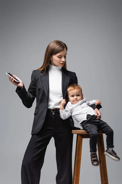Молодая бизнесвумен, держащая смартфон рядом с ребенком, сидящим на высоком стуле, изолированном на сером — стоковое фото