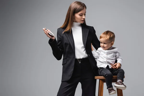 Woman in black suit standing with smartphone near son sitting on high stool isolated on grey — Photo de stock