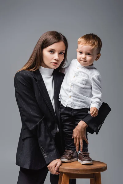Joven mujer mirando a cámara y abrazando hijo de pie en alto taburete aislado en gris - foto de stock