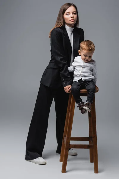 Full length view of woman in black suit standing near son on high stool on grey — стоковое фото