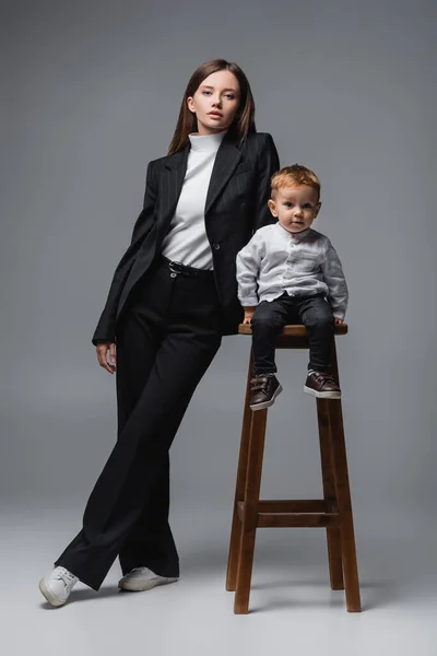 Bebé niño sentado en el taburete alto y mirando a la cámara cerca de mamá en traje negro en gris - foto de stock