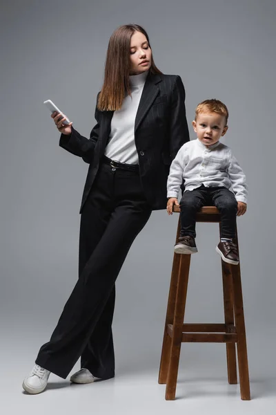 Ganzkörperansicht einer Frau im schwarzen Anzug, die ihr Smartphone in der Nähe ihres Sohnes hält, der auf einem hohen Schemel auf grau sitzt — Stockfoto