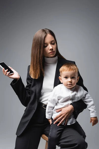 Frau hält Smartphone mit leerem Bildschirm und Kleinkindsohn isoliert auf grau — Stockfoto