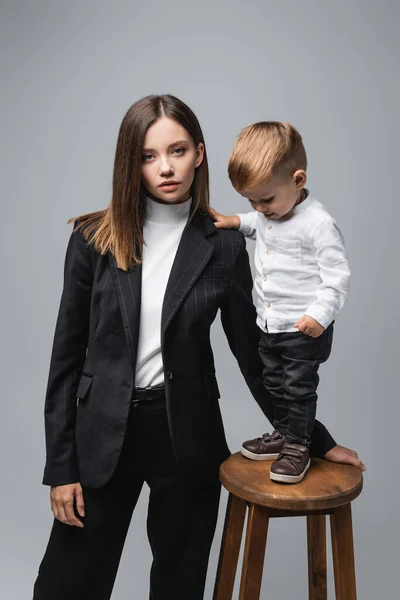 Bambino in piedi su sgabello alto vicino alla madre guardando la fotocamera isolata sul grigio — Foto stock