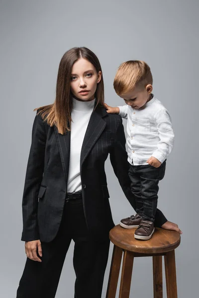 Pequeño niño de pie en el taburete alto cerca de la madre mirando a la cámara aislada en gris - foto de stock