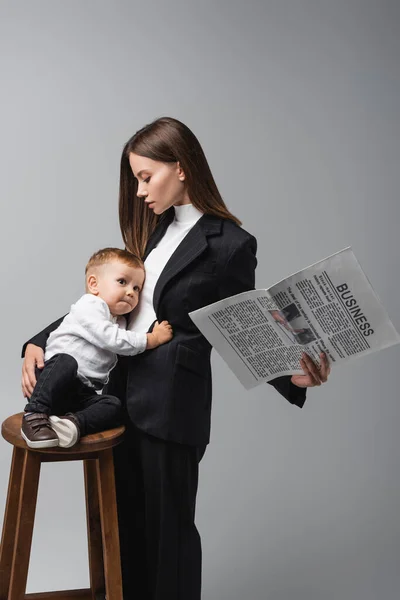 Женщина в черном костюме держит газету и обнимает маленького сына, сидящего на высоком стуле, изолированном на сером — стоковое фото
