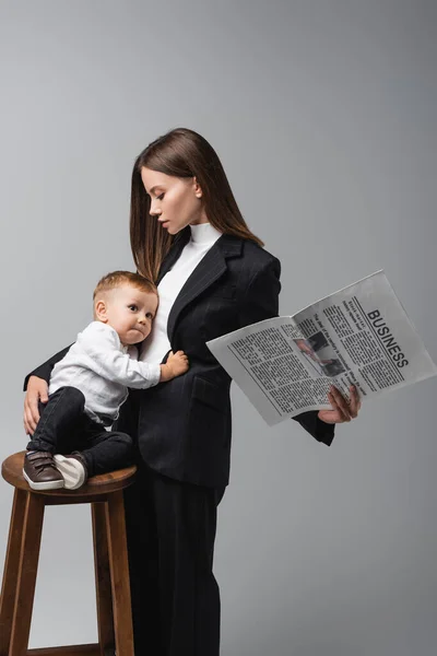 Woman in black suit holding newspaper and embracing little son sitting on high stool isolated on grey — Stock Photo