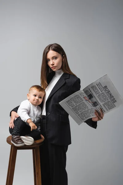 Mujer con negocio periódico abrazo hijo sentado en alto taburete aislado en gris - foto de stock