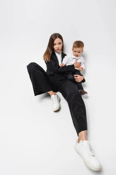 Giovane donna in completo nero seduta con piccolo figlio su bianco — Foto stock
