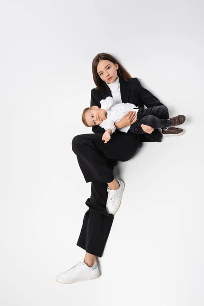 Vue grand angle de la jeune femme en costume noir assis avec petit enfant sur blanc — Photo de stock