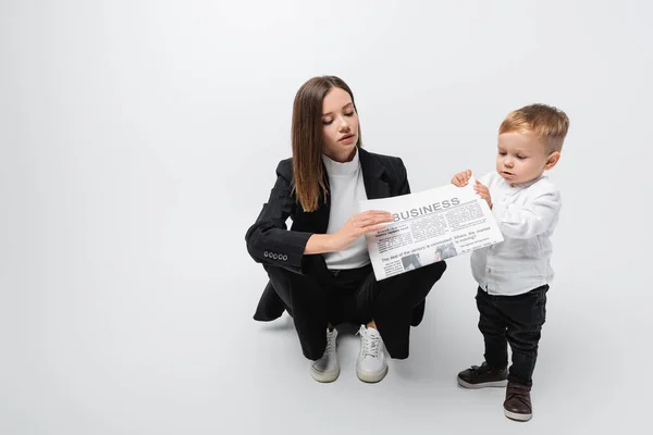 Woman in black suit holding newspaper near little son while sitting on grey — Stock Photo
