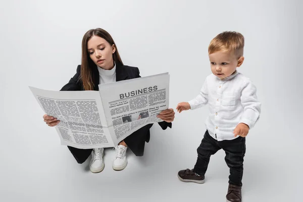 Businesswoman sitting and reading newspaper near toddler son on grey — стоковое фото