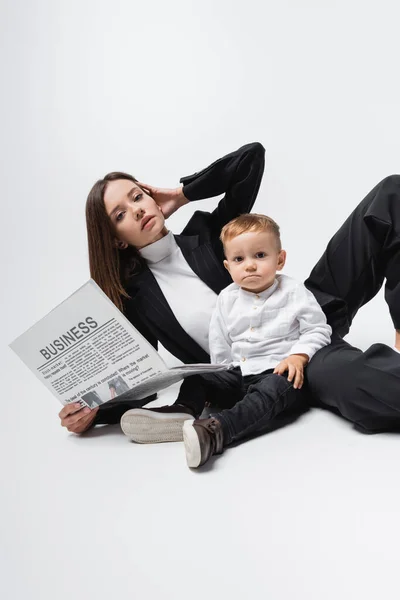 Mujer de negocios con periódico mirando a la cámara cerca de hijo pequeño en blanco - foto de stock