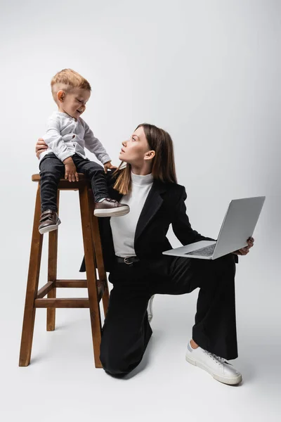Woman with laptop hugging toddler son sitting on high stool on grey — Photo de stock