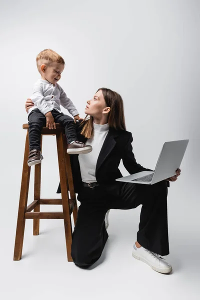 Woman with laptop hugging toddler son sitting on high stool on grey — Stock Photo