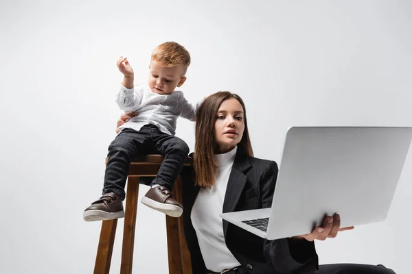 Empresária olhando para laptop perto do filho sentado em banquinho alto isolado em cinza — Fotografia de Stock