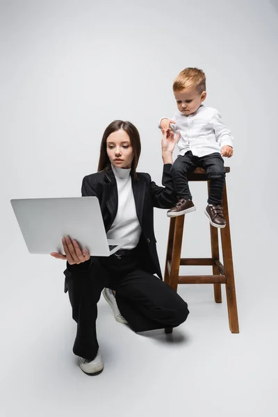 Businesswoman with laptop holding hands with little son sitting on high stool on grey — Stock Photo