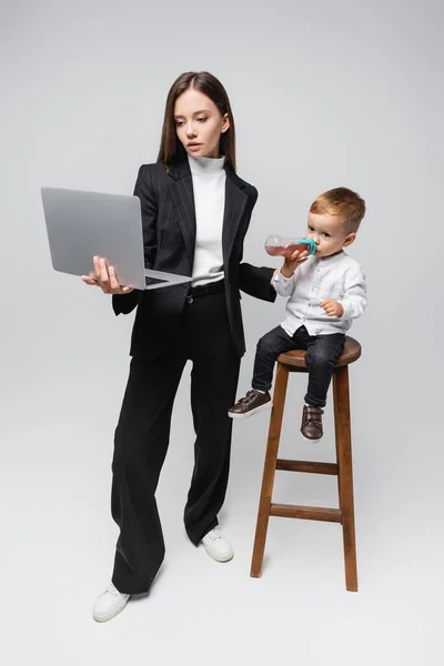 Kleinkind trinkt aus Babyflasche, während es auf hohem Hocker neben Mama sitzt, Laptop auf grau — Stockfoto