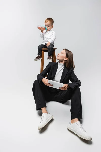 Woman with digital tablet looking at son drinking from baby bottle on white — стоковое фото