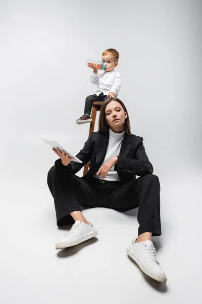 Woman in black suit sitting with digital tablet near son on high stool drinking from baby bottle on grey — Stock Photo