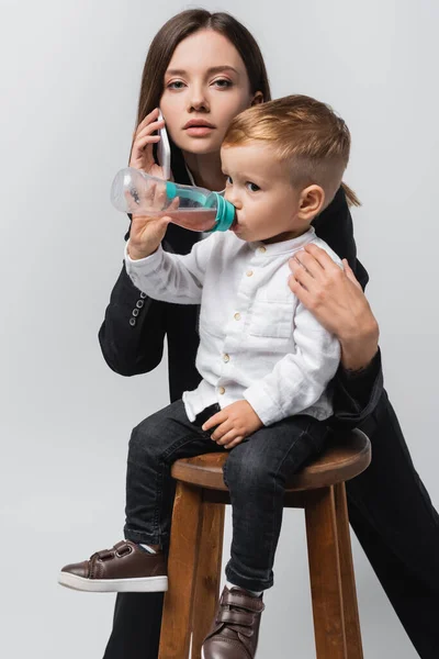 Young woman talking on smartphone near son drinking from baby bottle isolated on grey — Stockfoto