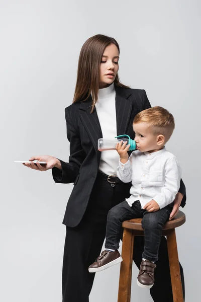 Женщина в черном костюме держит смартфон рядом с сыном, пьющим из детской бутылочки, изолированной на сером — стоковое фото