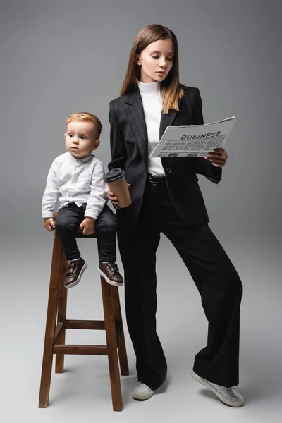 Niño sentado en el taburete alto cerca de la madre con café para ir y periódico de negocios en gris - foto de stock