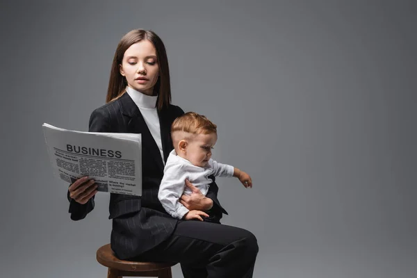 Woman in black suit reading business newspaper while sitting with son on high stool isolated on grey — Stock Photo