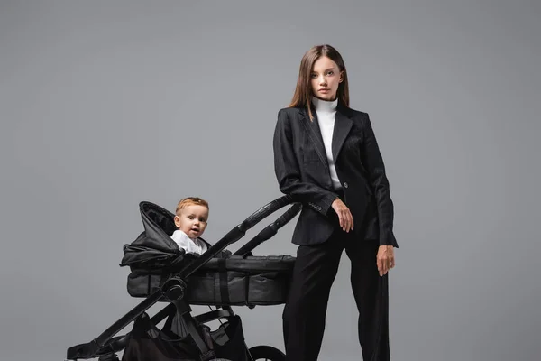 Mujer de negocios en traje mirando a la cámara cerca de hijo en cochecito negro aislado en gris - foto de stock