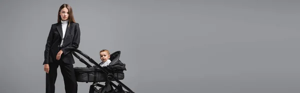 Young businesswoman looking at camera near son sitting in black pram isolated on grey, banner — Photo de stock