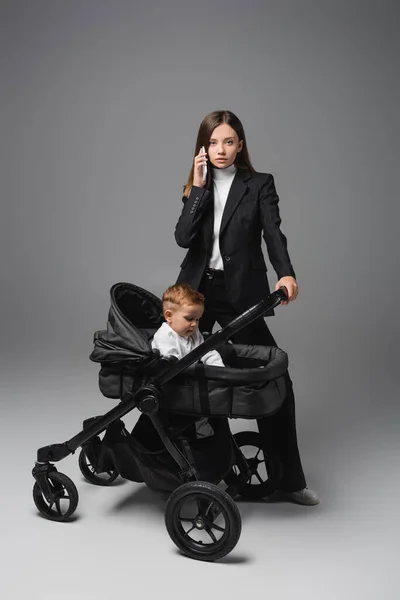 Young woman calling on mobile phone near son in baby carriage on grey — Foto stock