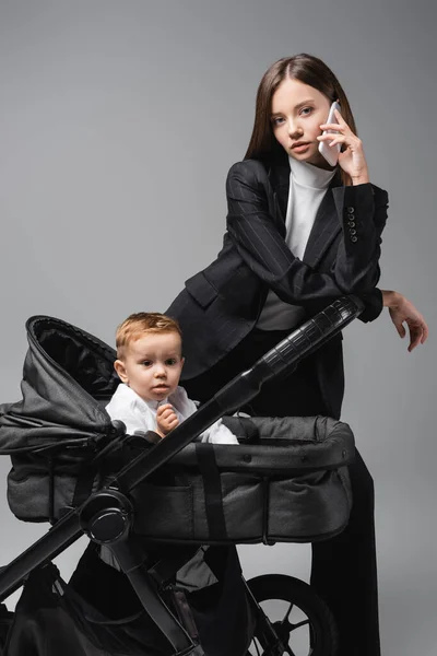 Mujer en traje hablando en el teléfono móvil cerca de hijo pequeño en carro de bebé aislado en gris - foto de stock