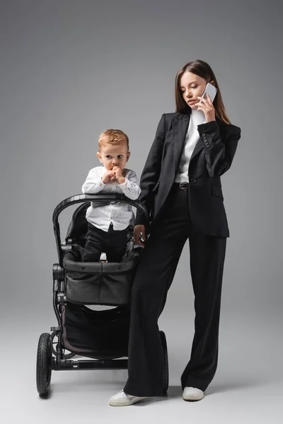 Businesswoman standing near little son in pram during conversation on smartphone on grey — Stockfoto