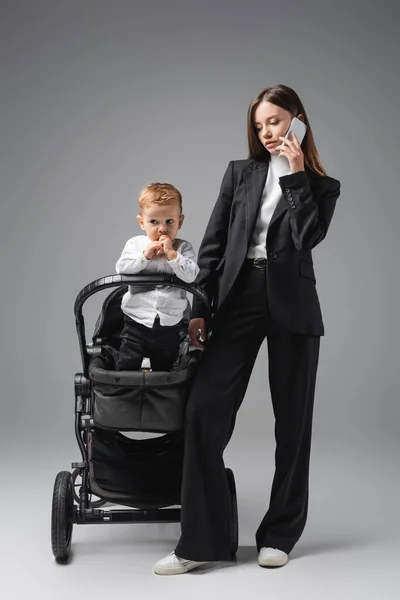 Businesswoman standing near little son in pram during conversation on smartphone on grey — Stock Photo